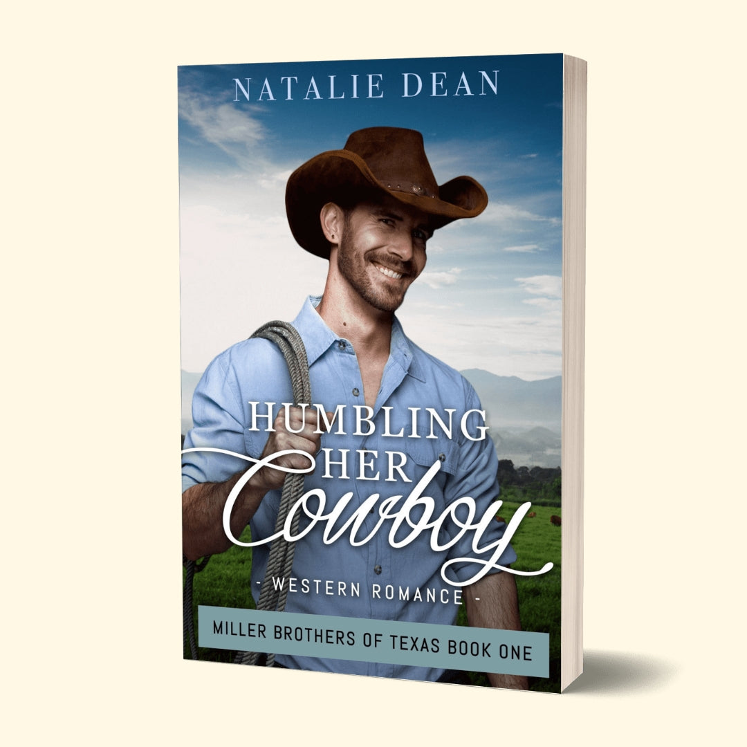 3D mockup of the book “Humbling Her Cowboy” by Natalie Dean. The cover features a smiling cowboy in a brown hat and blue shirt holding a rope, standing against a scenic ranch backdrop with green fields and blue mountains under a partly cloudy sky. Subtitle reads “Western Romance,” and the bottom banner says “Miller Brothers of Texas Book One.”