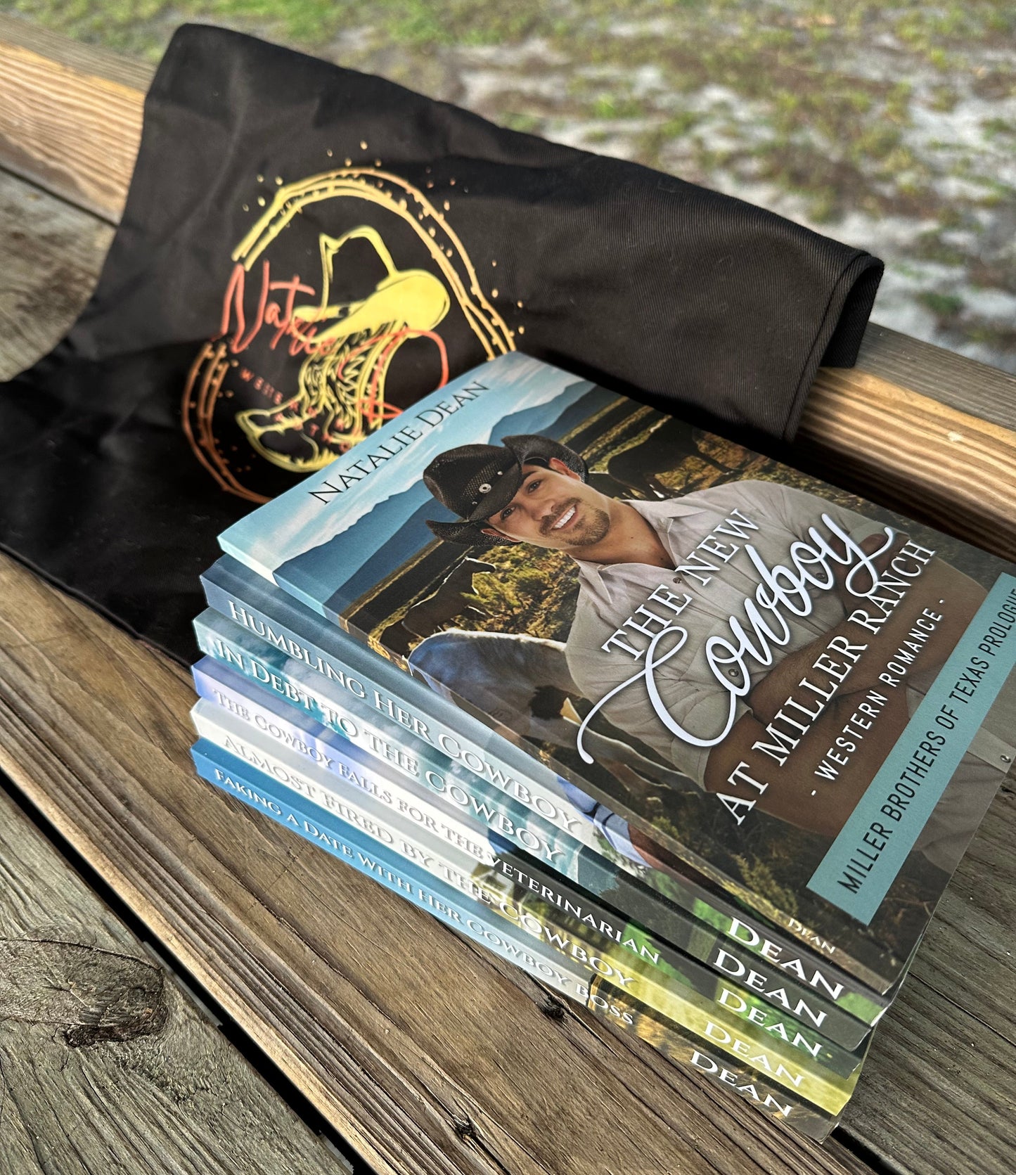 Stack of all six Miller Brothers of Texas books on a wooden surface with a black tote bag featuring the Natalie Dean logo. The logo on the bag has a line image of a pair of cowboy boots with a cowboy hat sitting on top, along with Natalie Dean written in a pretty script. 