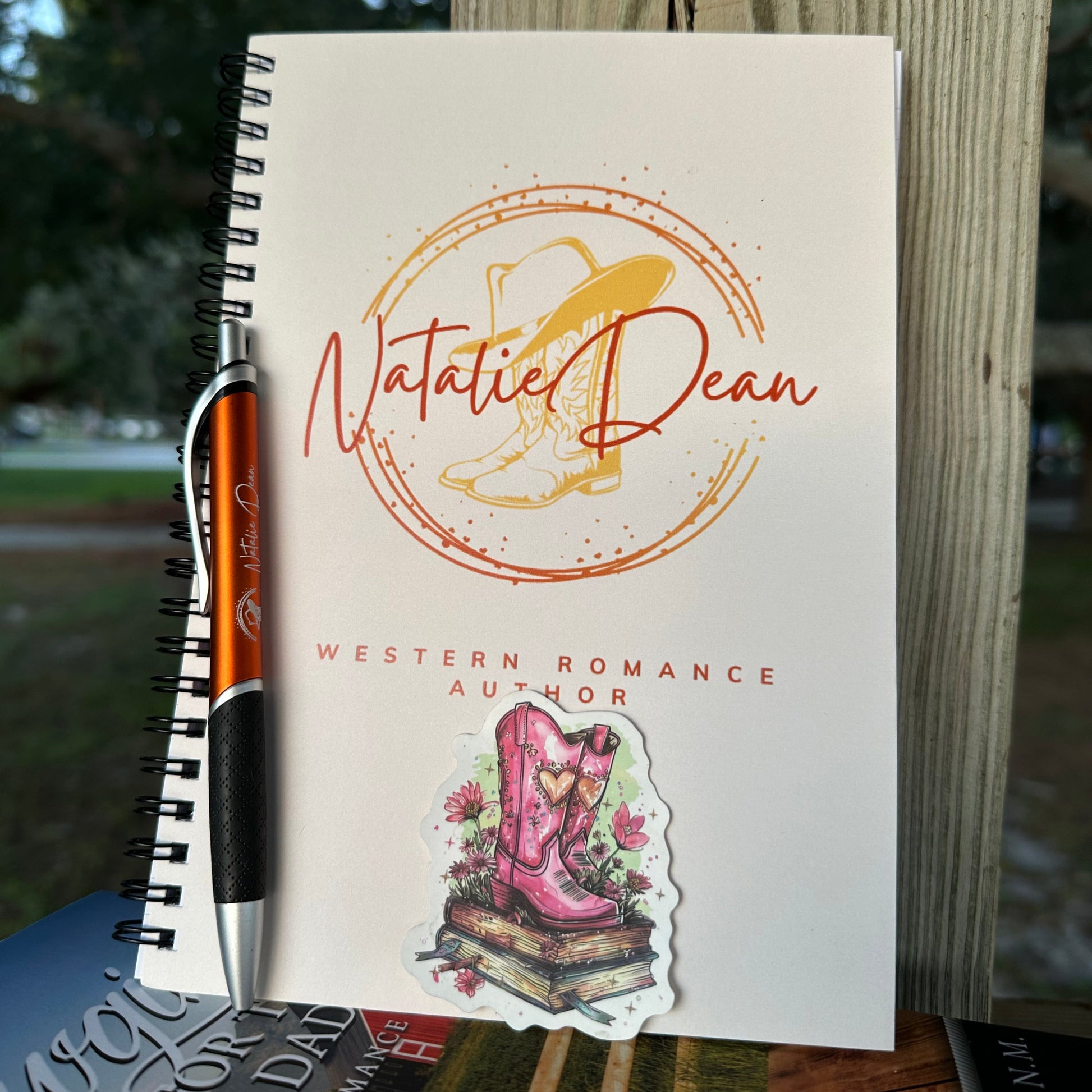 Notebook with the Natalie Dean logo (Natalie Dean signature, cowboy boots and a cowboy hat on top). A burnt orange ballpoint pen with the Natalie Dean logo on it. a sticker with pretty cowgirl boots, flowers both sitting on top of a stack of books.