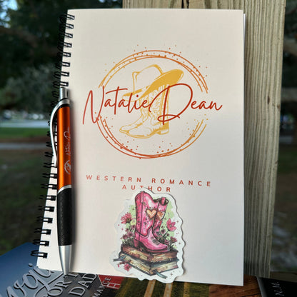 Notebook with the Natalie Dean logo (Natalie Dean signature, cowboy boots and a cowboy hat on top). A burnt orange ballpoint pen with the Natalie Dean logo on it. a sticker with pretty cowgirl boots, flowers both sitting on top of a stack of books.