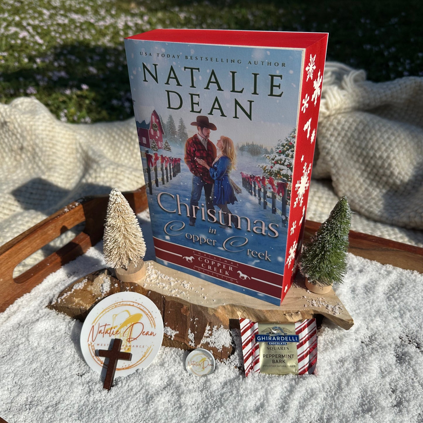 “Standing display of the Christmas in Copper Creek paperback with red sprayed edges and winter stencils, surrounded by faux snow, 2 mini Christmas trees, peppermint bark, a Natalie Dean sticker, and a wooden cross.”