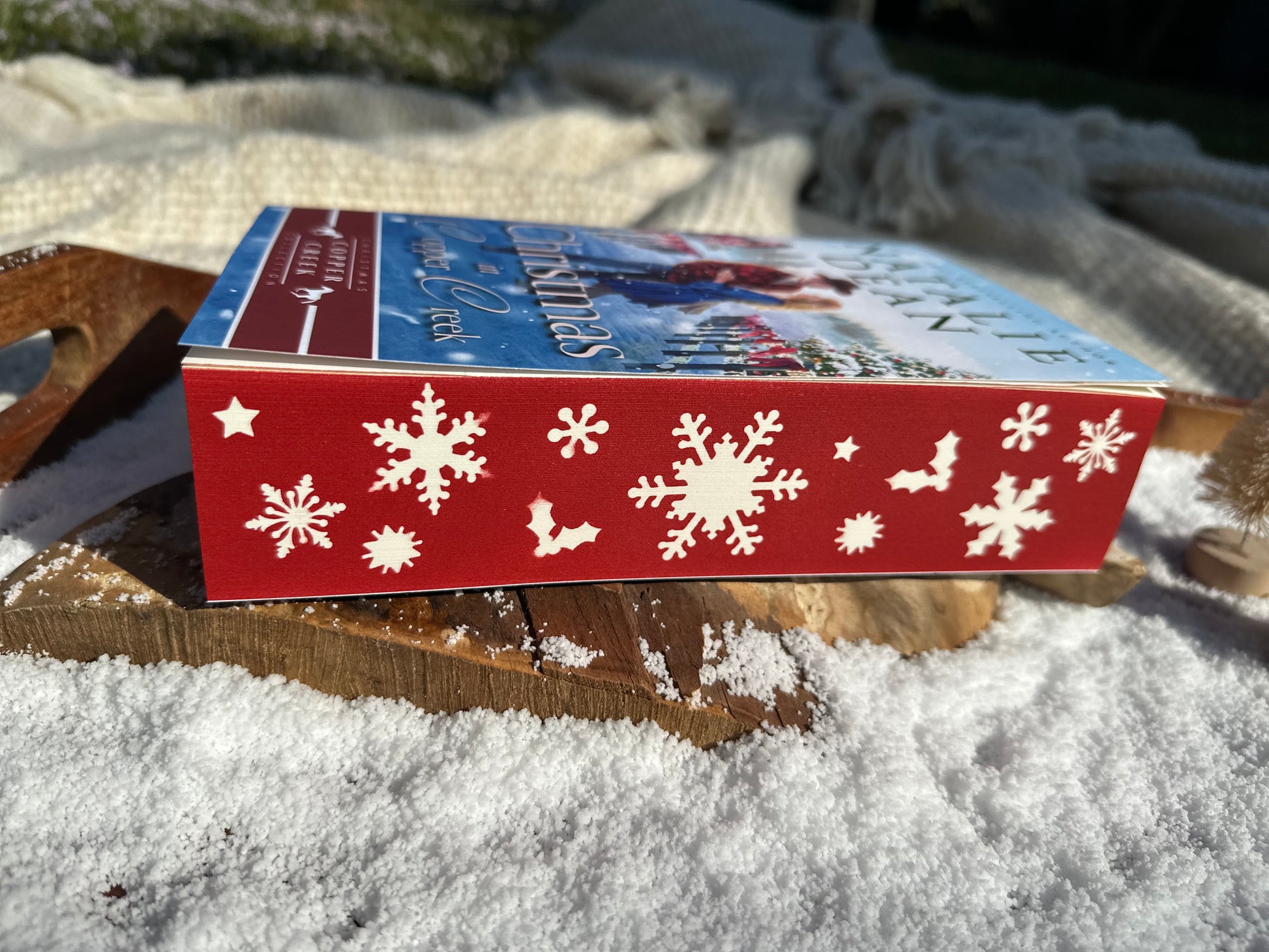 Side view of the Christmas in Copper Creek paperback showing red sprayed edges decorated with white stenciled snowflakes, holly leaves, and stars, placed on faux snow with a rustic wood background.

