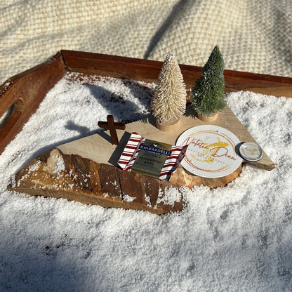 “Flat-lay display of the Christmas in Copper Creek book box items arranged on faux snow, including mini Christmas trees, a wood slice, peppermint bark chocolate, a Natalie Dean sticker, a wooden cross, and a glass-dome magnet.”